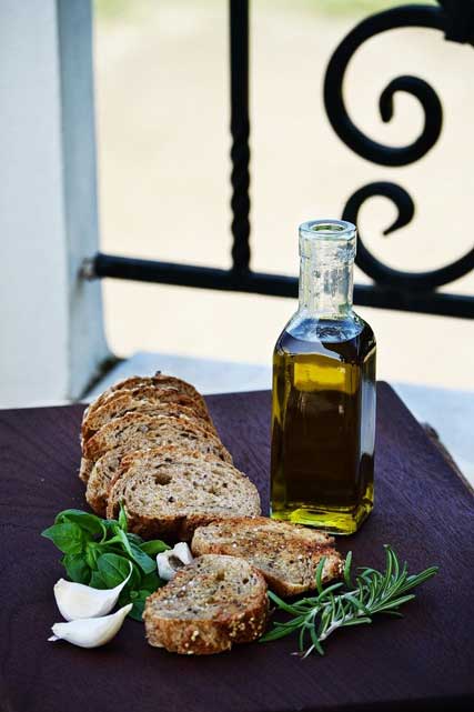 A small bottle of olive oil with next to it some garlic and sliced bread, all decorated with rozmarin